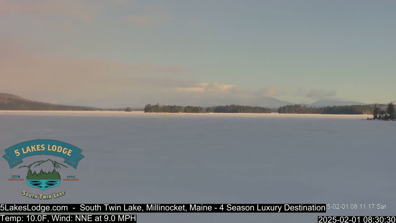 A snow-covered lake stretches to a treeline and distant mountains under a mostly clear, pale sky.