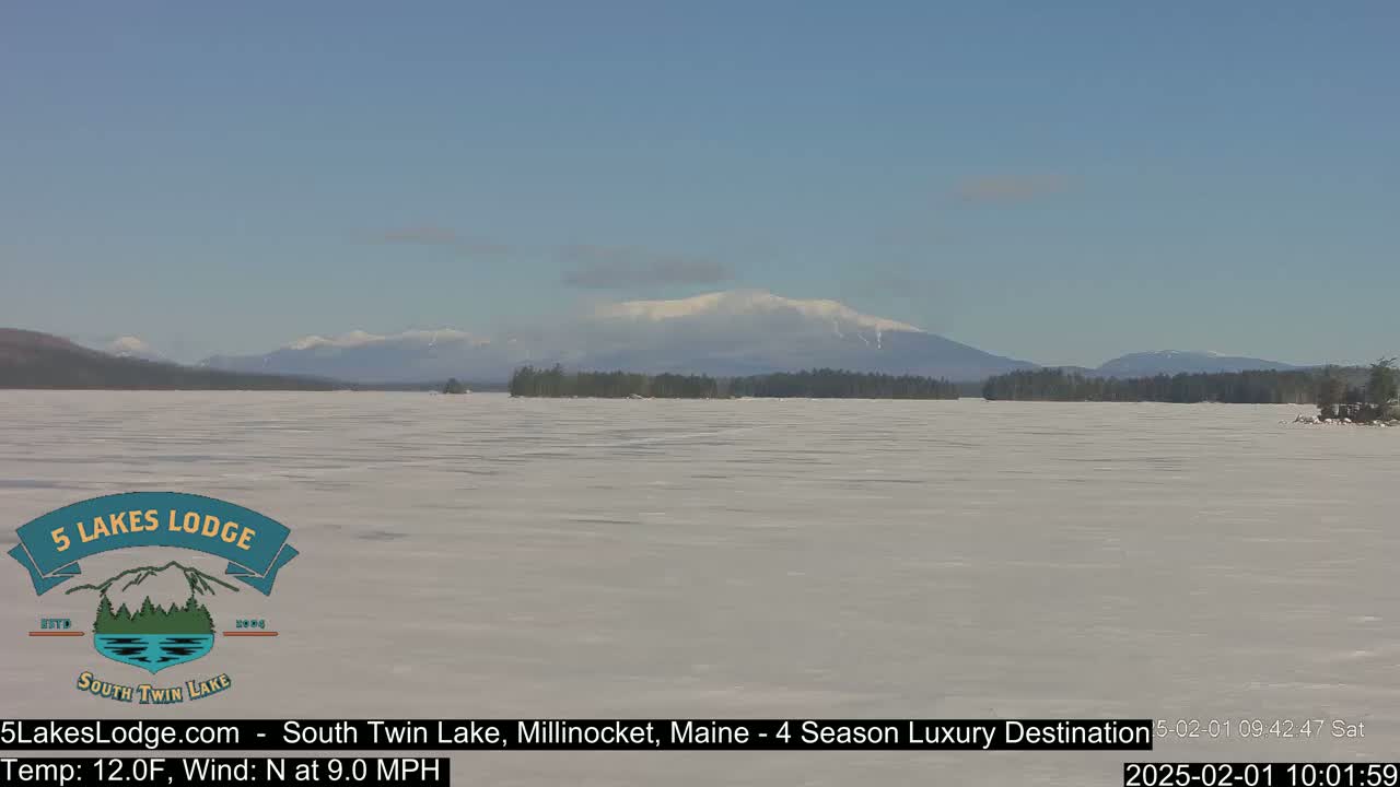 A frozen lake stretches out to snow-covered mountains under a mostly clear, bright sky.