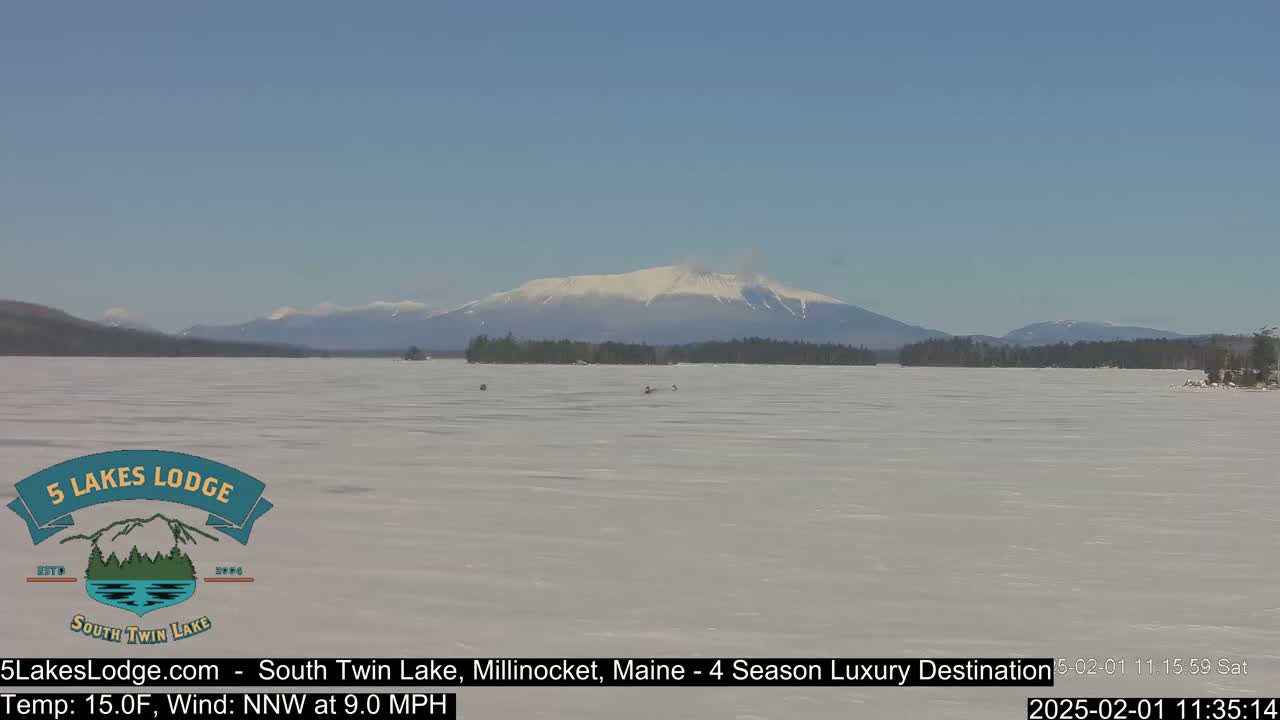 A snow-covered lake stretches towards a distant, snow-capped mountain range under a clear blue sky.