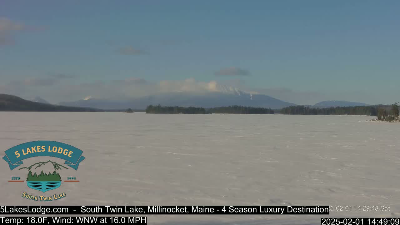 A frozen lake stretches to snow-capped mountains under a mostly clear, light blue sky.