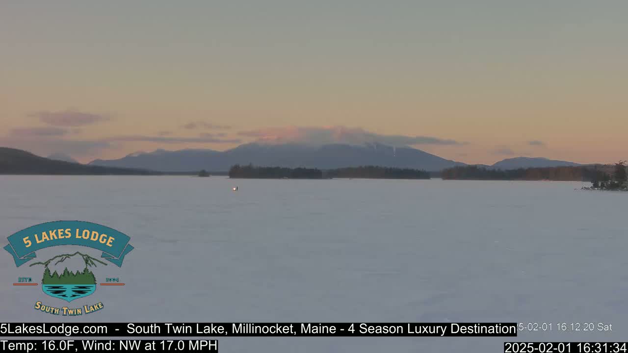 A snow-covered lake stretches to distant mountains under a pale pink and gray sunset.