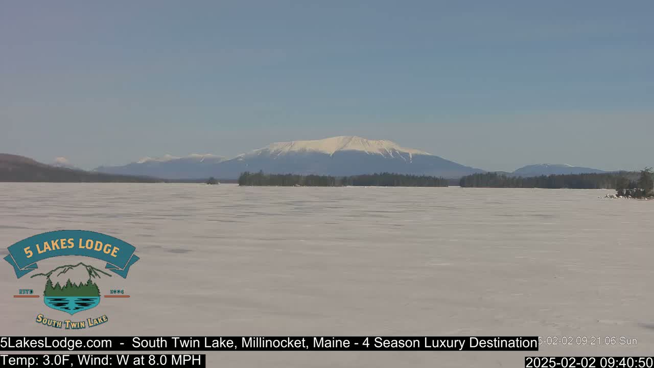 A snow-covered lake stretches toward a distant, snow-capped mountain range under a clear blue sky.