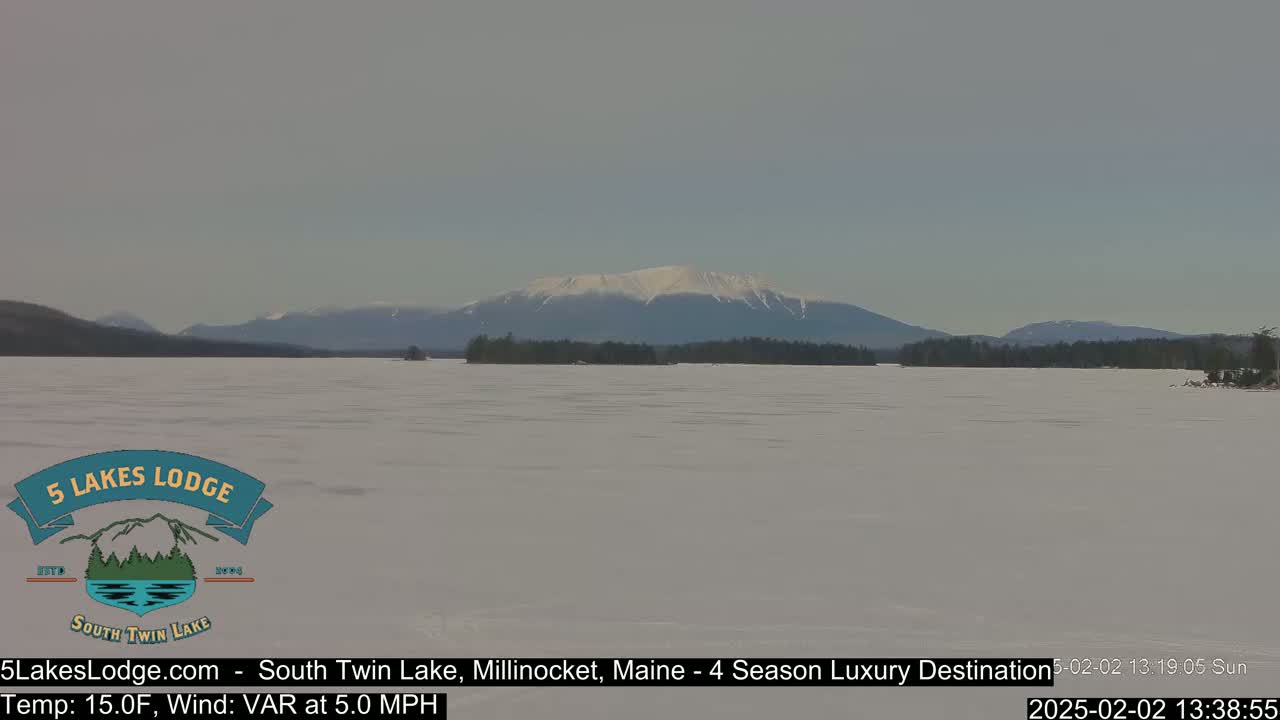 A snow-covered lake stretches to a distant mountain range partially covered in snow under a clear, light gray sky.