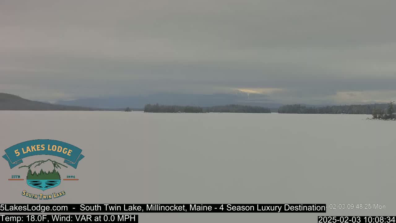 A snow-covered lake stretches to distant, snow-capped mountains under an overcast sky.