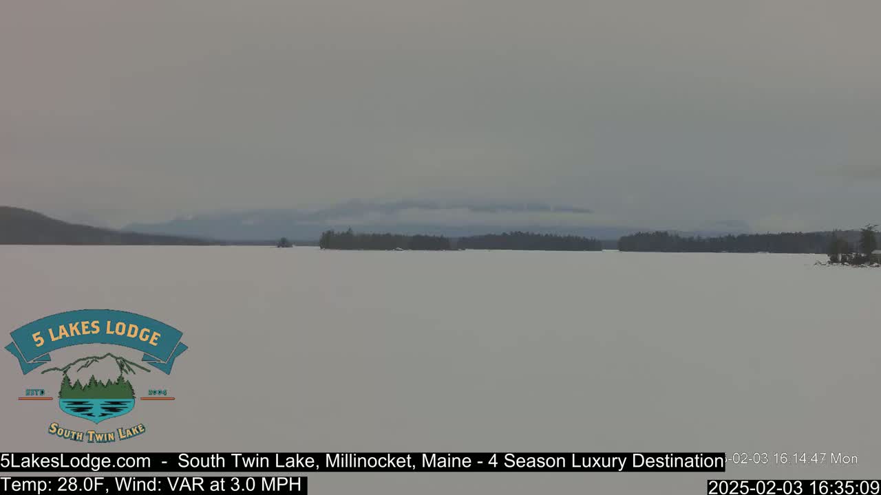 A snow-covered lake stretches to a distant, tree-lined shore under an overcast sky.