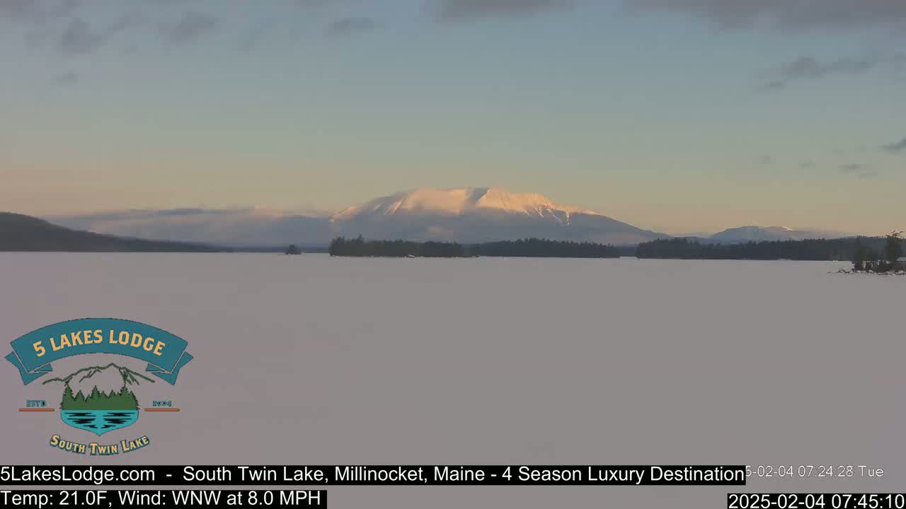 A snow-covered lake stretches to a distant, snow-capped mountain range under a pale, partly cloudy sky.