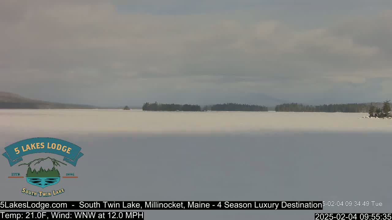 A mostly cloudy day shows a frozen lake with a distant treeline under a pale sky.