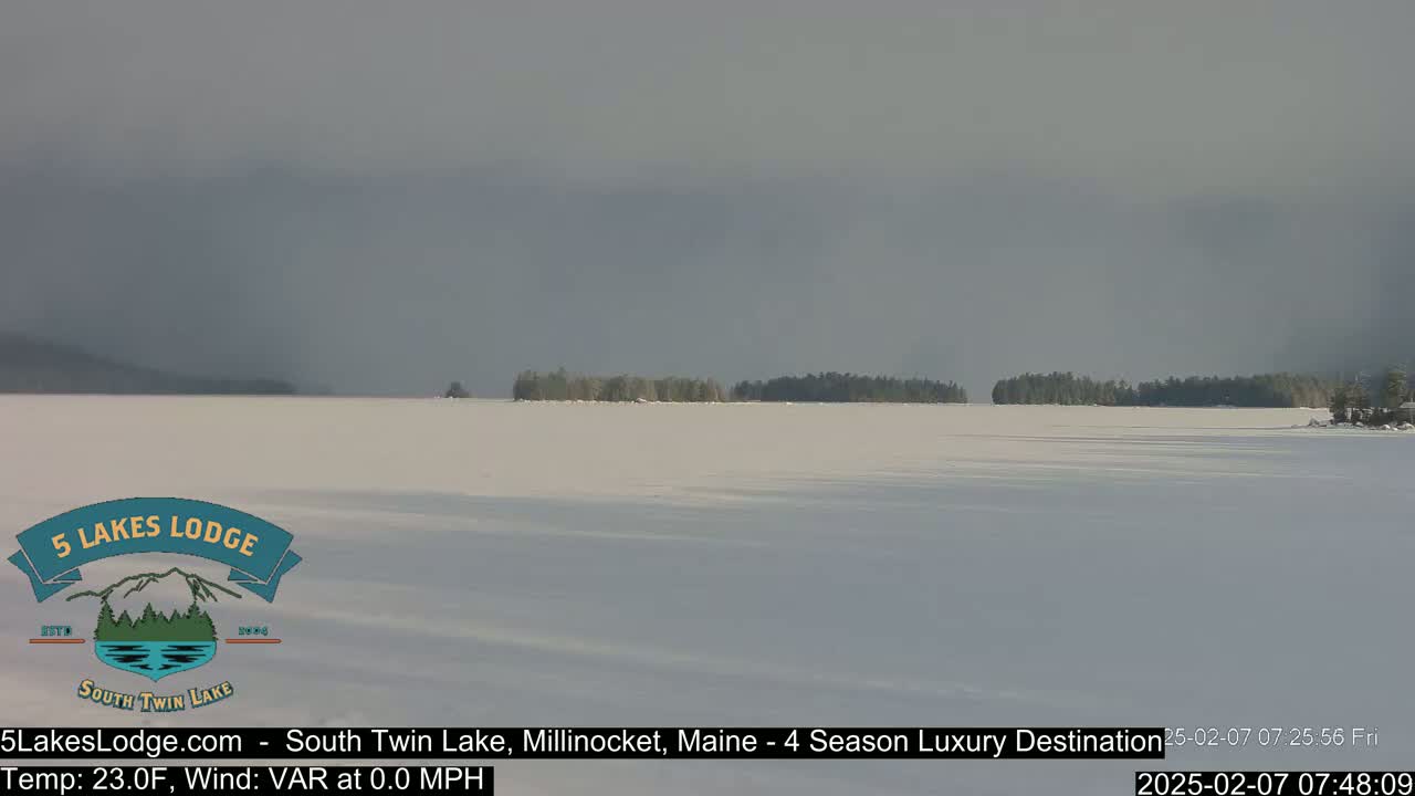 A snow-covered lake stretches to a distant treeline under a cloudy sky.