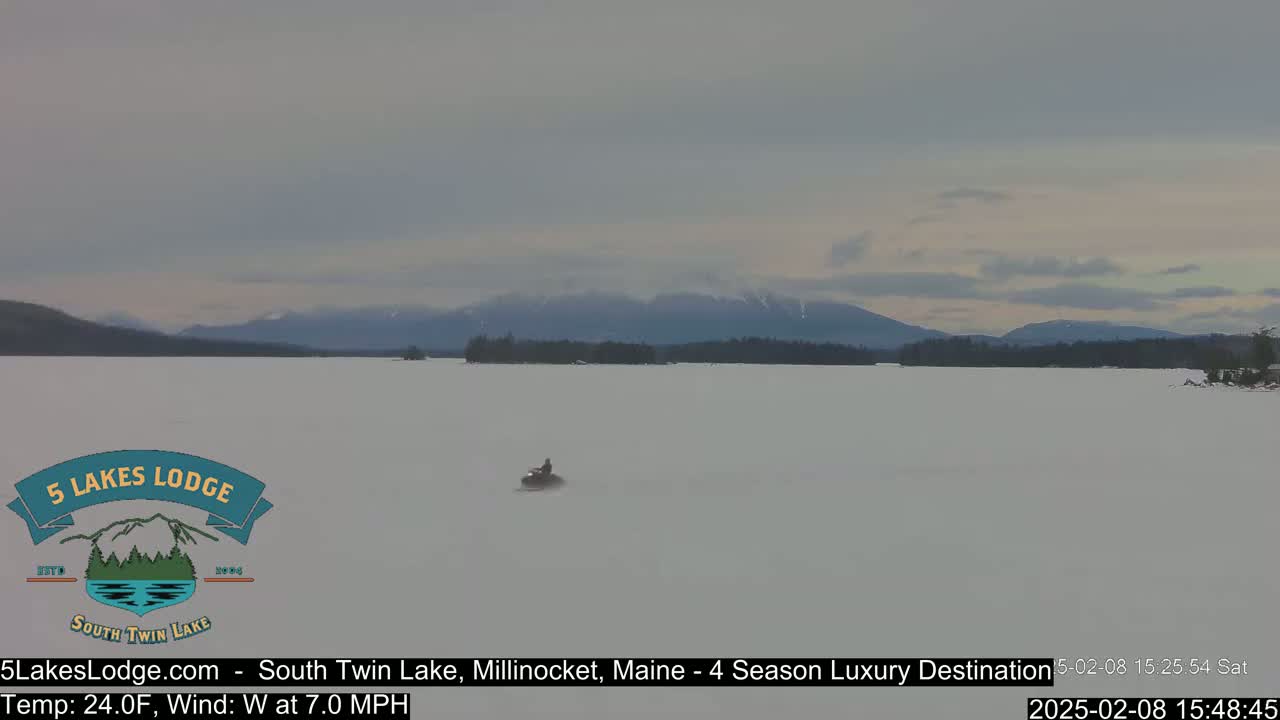 A snowmobile travels across a frozen lake under a cloudy sky, with distant mountains visible on the horizon.