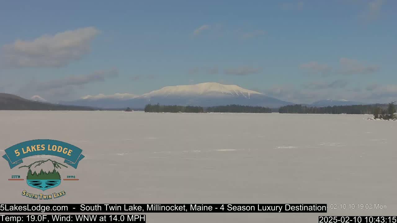 A snow-covered lake stretches towards a snow-capped mountain range under a partly cloudy sky.