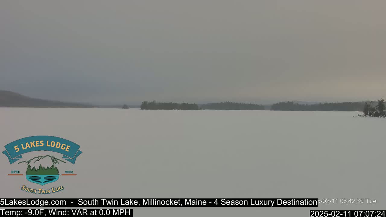 A snow-covered lake stretches to a distant, tree-lined shore under a gray, overcast sky.
