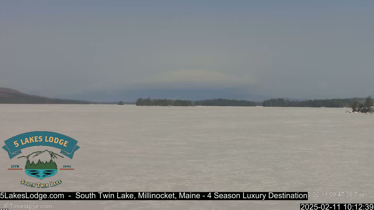 A mostly clear day reveals a frozen lake with a distant shoreline of trees under a pale sky.