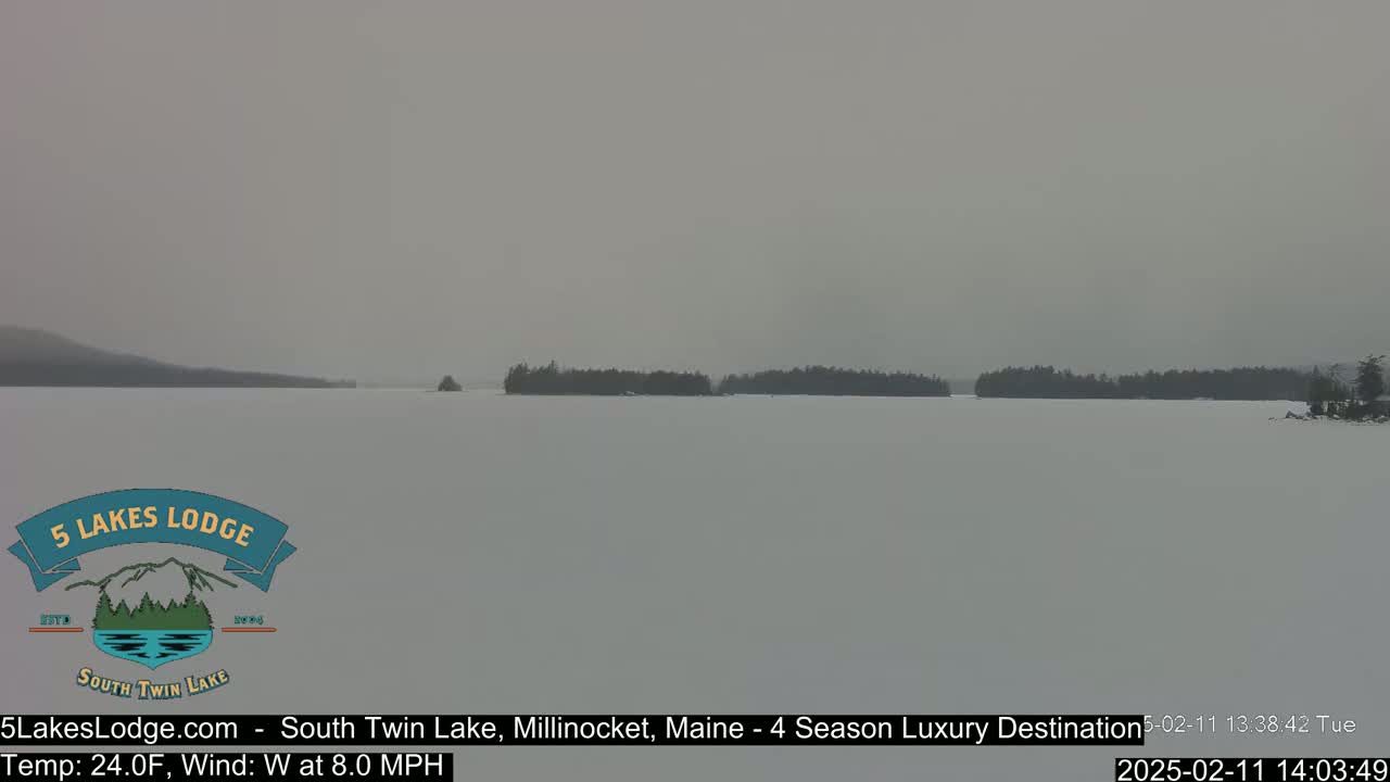 A snow-covered lake is visible under a hazy, overcast sky, with dark tree lines in the far distance.