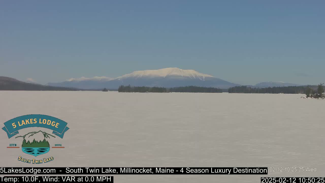 A snow-covered lake stretches out to a snow-capped mountain range under a clear blue sky.
