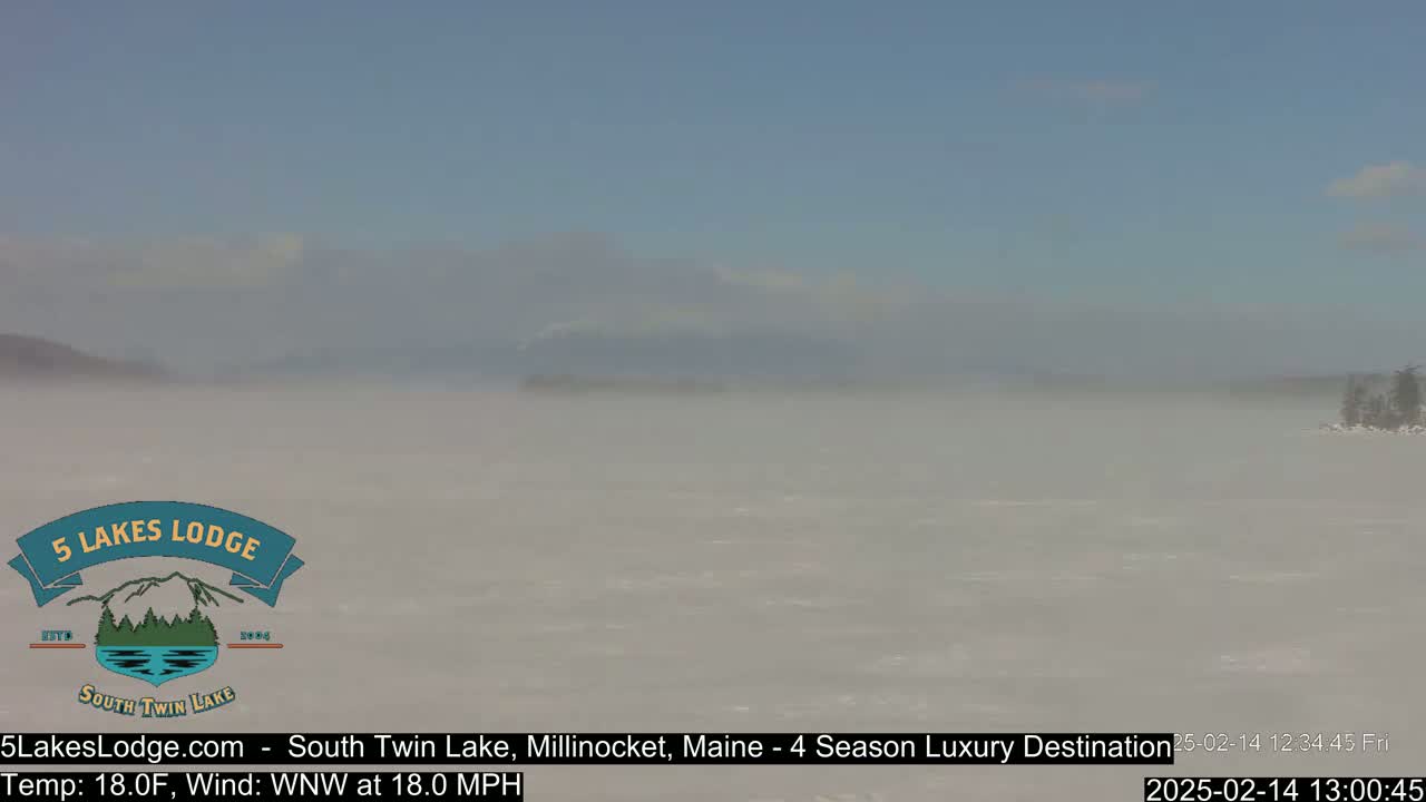 A hazy, snowy expanse of lake stretches to distant, indistinct mountains under a pale sky, with a strong wind blowing.