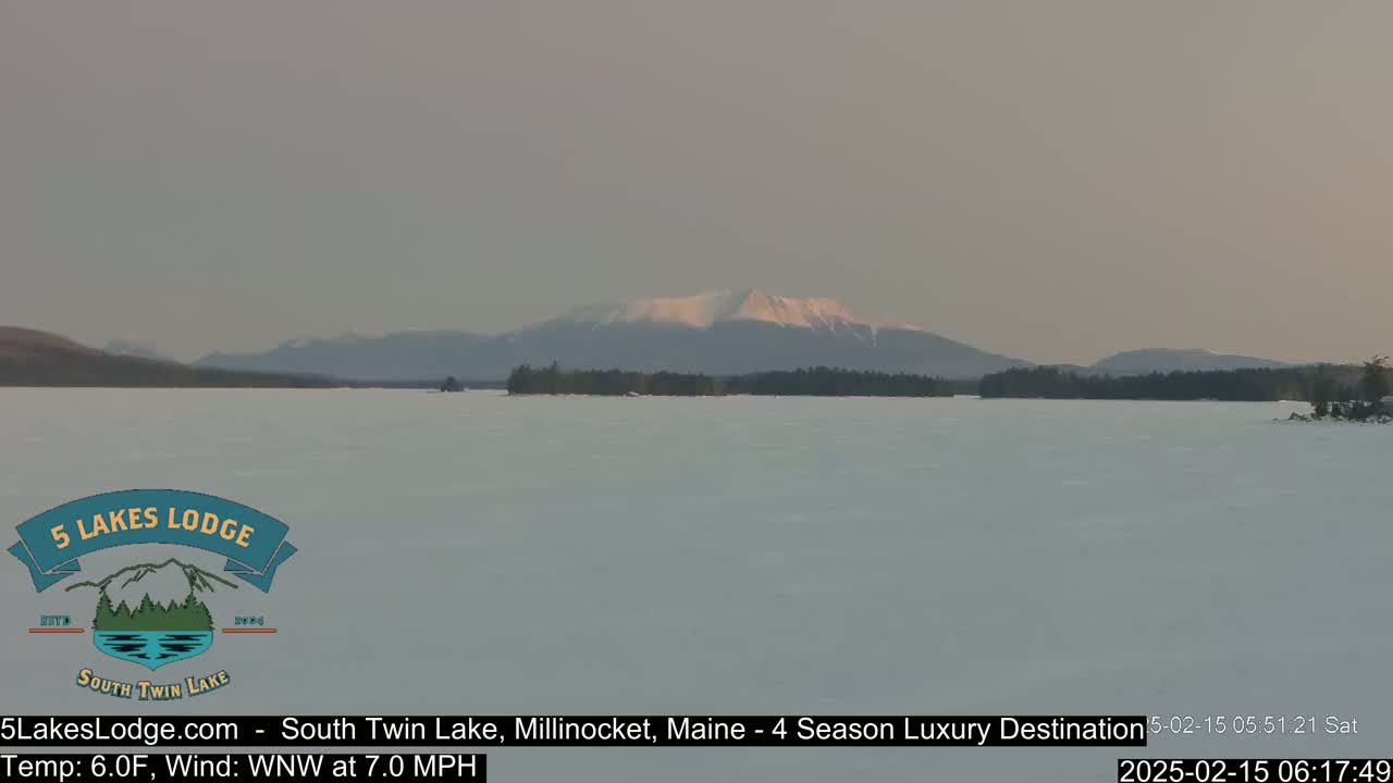 A snow-covered lake stretches to a snow-capped mountain range under a hazy sky.