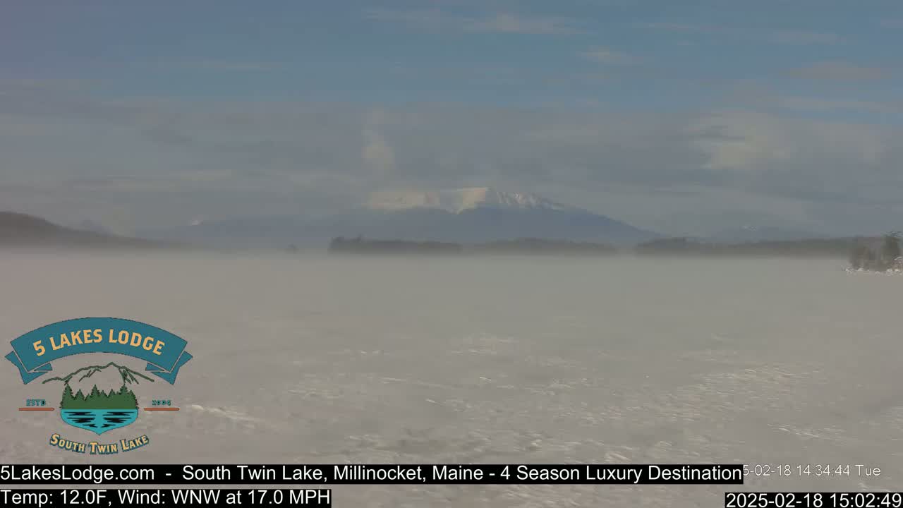 A foggy lake stretches toward snow-capped mountains under a mostly cloudy sky.