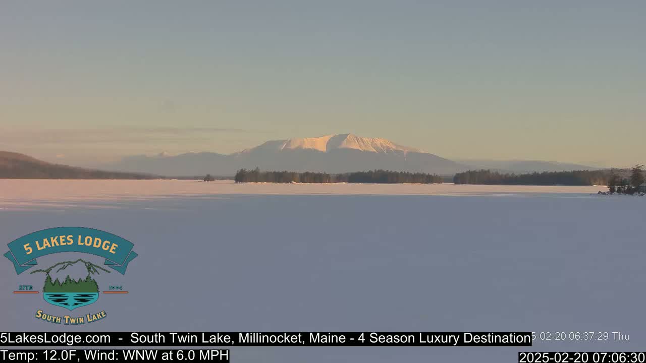 A snow-covered lake stretches towards a snow-capped mountain range under a pale, clear sky.