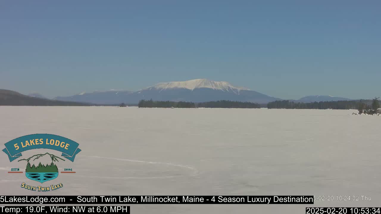A snow-covered lake stretches to a distant, snow-capped mountain range under a clear blue sky.