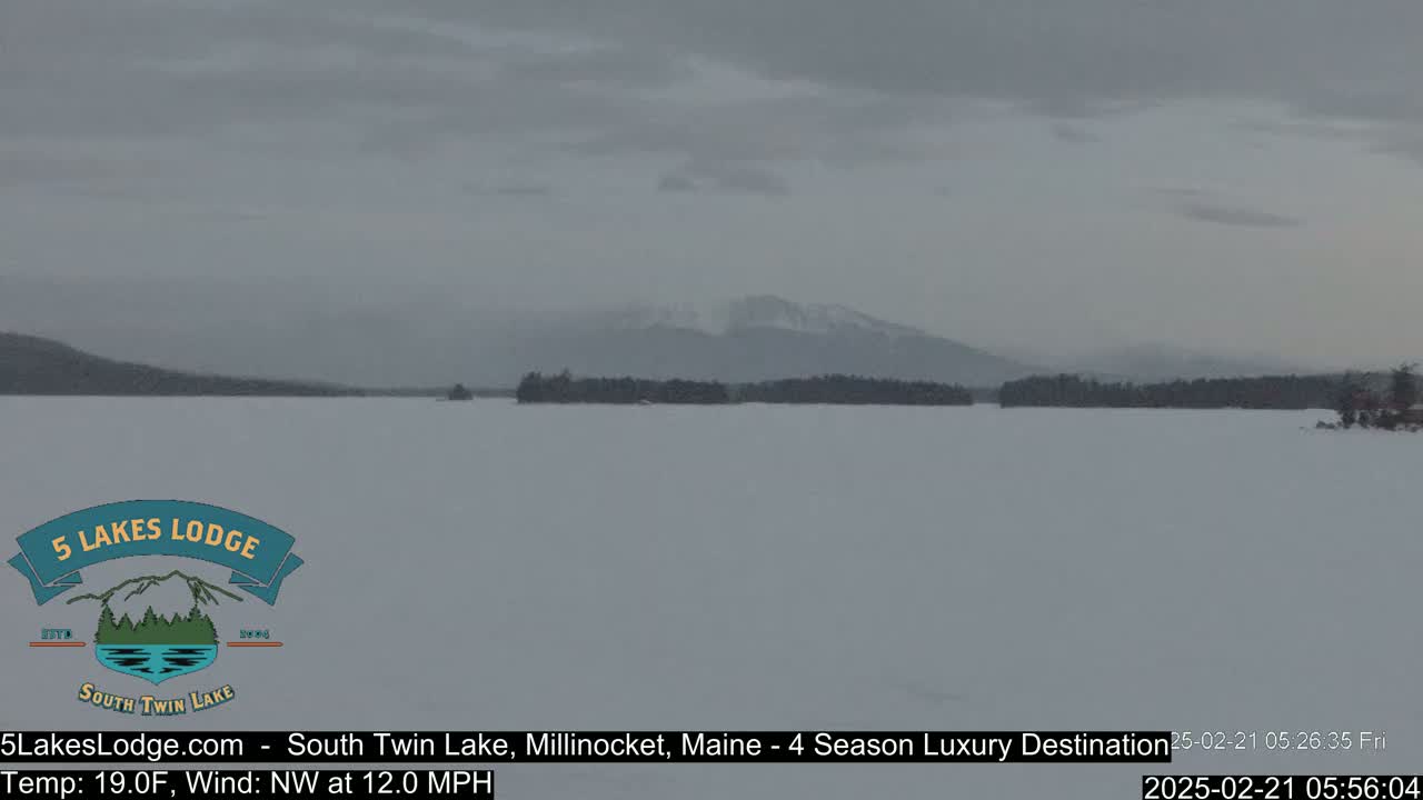 A snow-covered lake stretches to a distant, tree-lined shore beneath a gray, overcast sky, with mountains barely visible in the background.