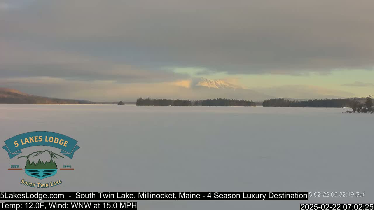 A snow-covered lake stretches to a distant mountain range under a cloudy, but brightening sky.