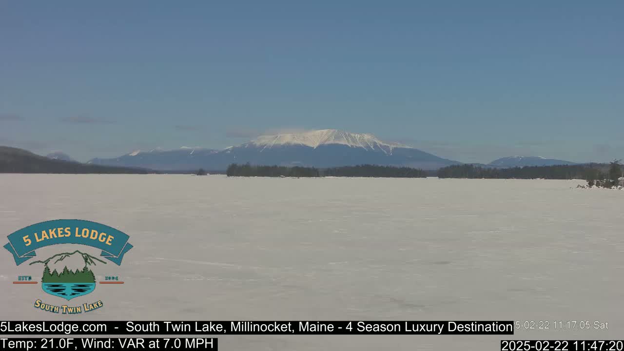 A snow-covered lake stretches to a distant, snow-capped mountain range under a clear blue sky.