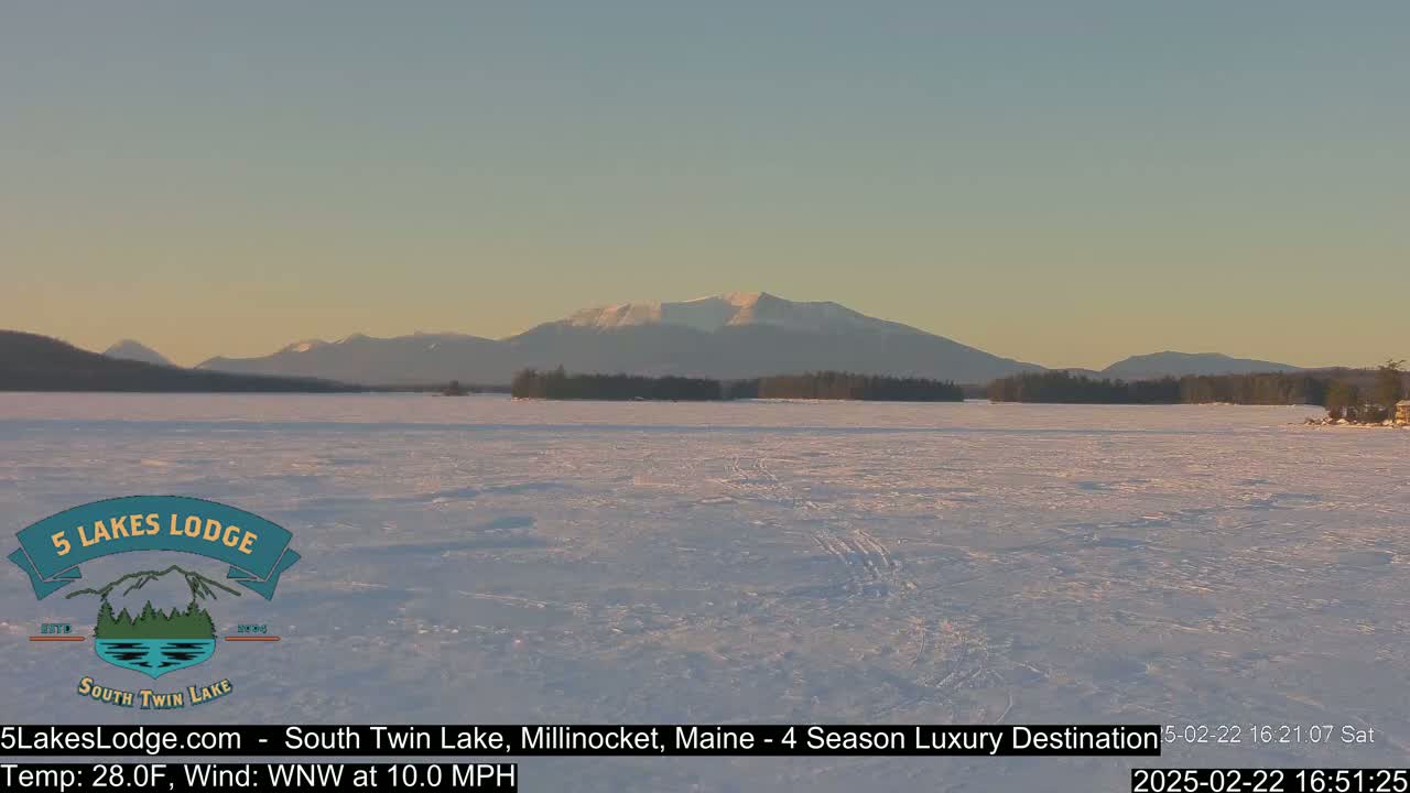 A snow-covered lake stretches to a mountain range under a clear, pale sky.