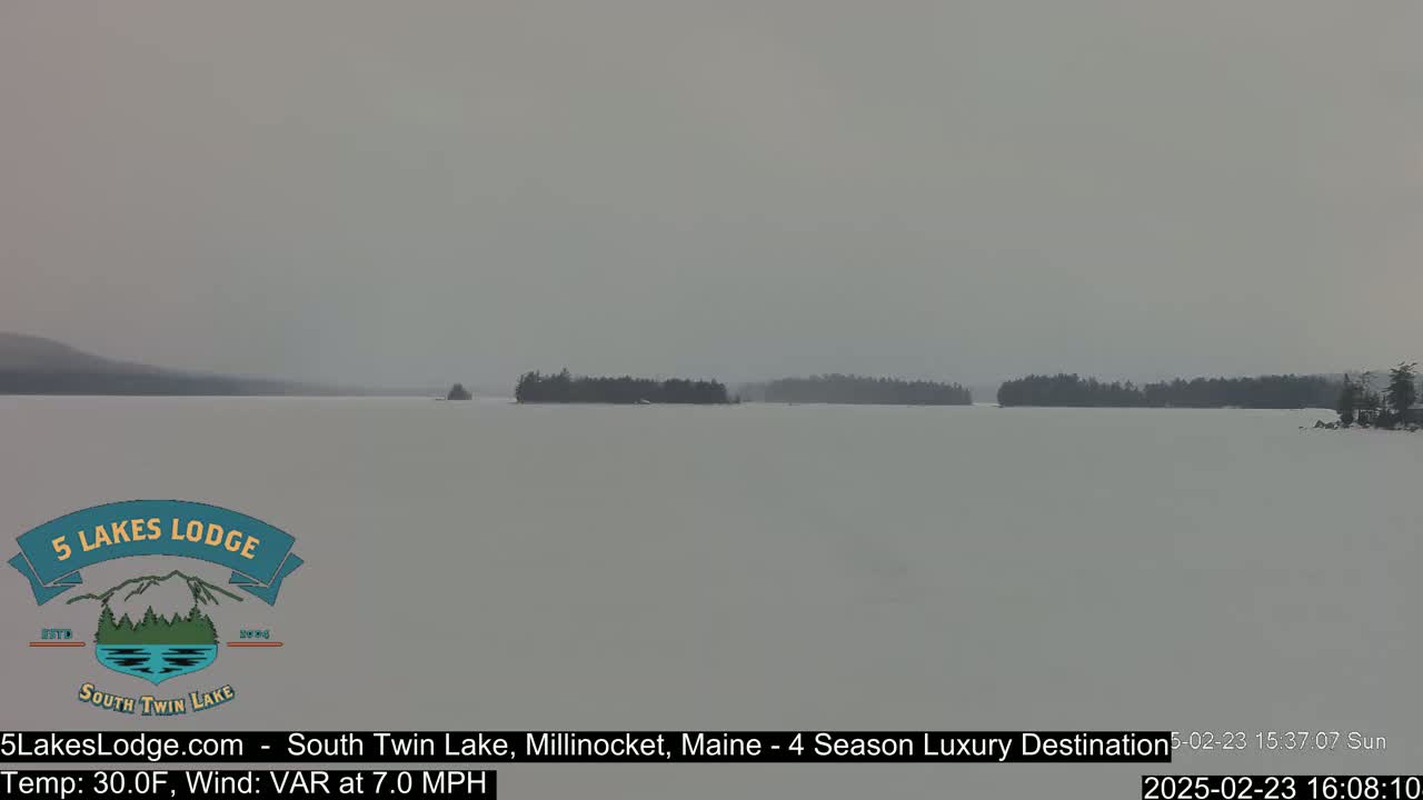 A snow-covered lake stretches to a distant shoreline of dark trees under a hazy, gray sky.