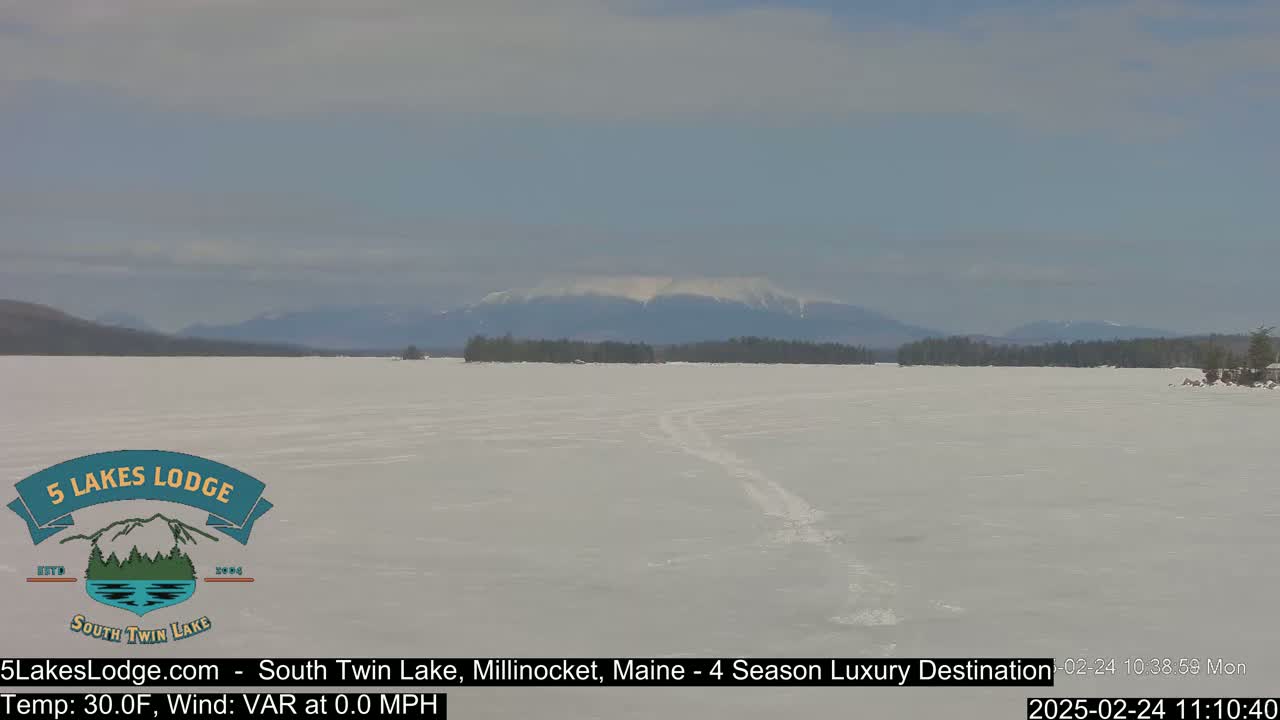 A snow-covered frozen lake stretches to a distant mountain range under a mostly clear sky.