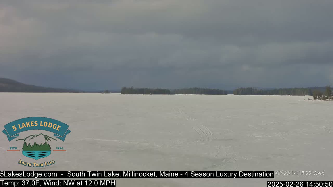 A snow-covered lake stretches to a treeline under a mostly cloudy sky.