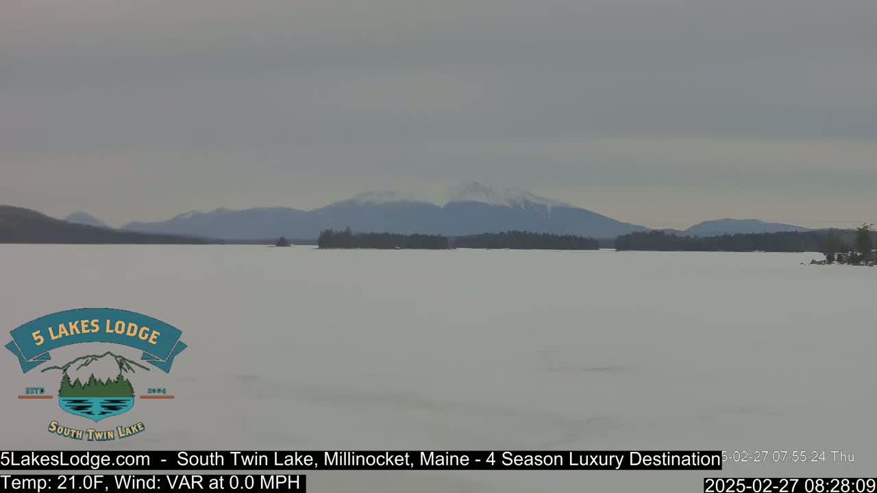 A snow-covered lake stretches out to a distant mountain range under a cloudy sky.
