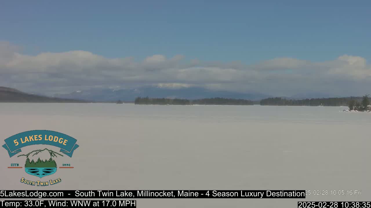 A frozen lake stretches to a distant, tree-lined shoreline under a partly cloudy sky.