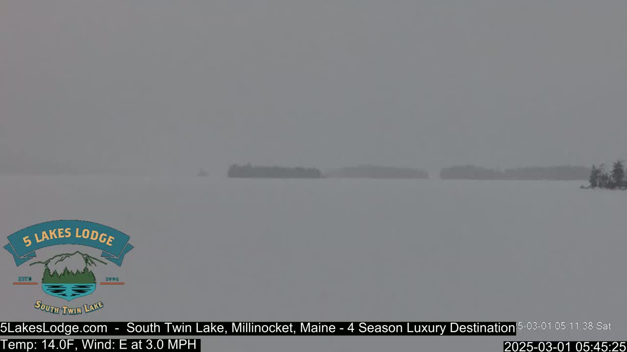 A snow-covered lake with distant, barely visible tree lines is seen under heavy snowfall.