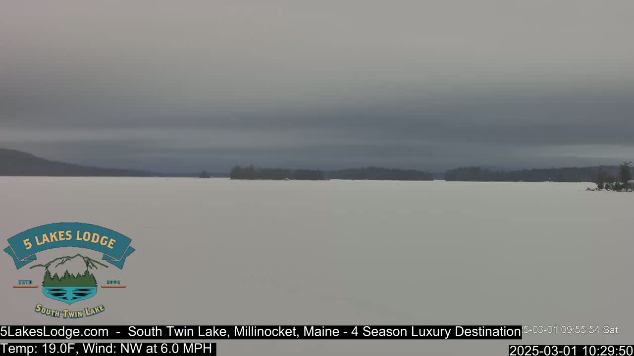 A snow-covered lake stretches to a distant, tree-lined shore under an overcast sky.