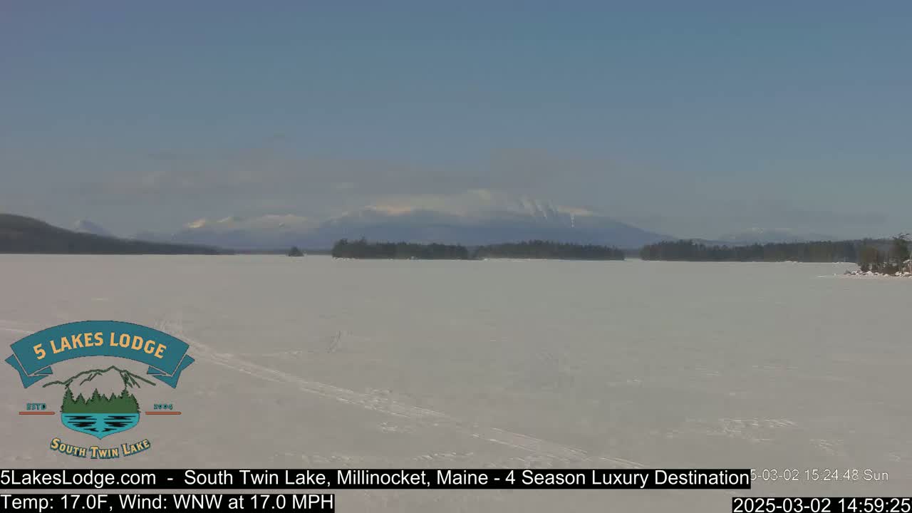 A frozen lake stretches towards snow-capped mountains under a clear blue sky, with a light wind blowing.