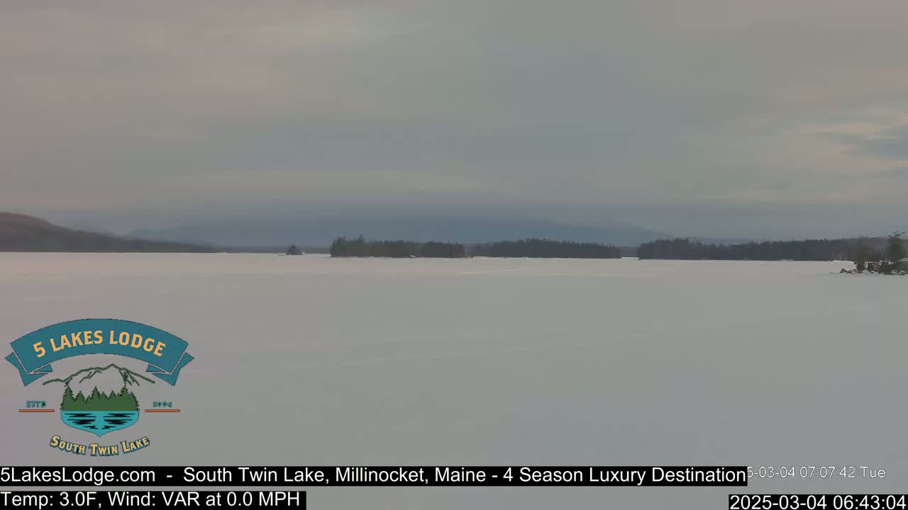 A snow-covered lake stretches to a distant, tree-lined shore under a cloudy sky.