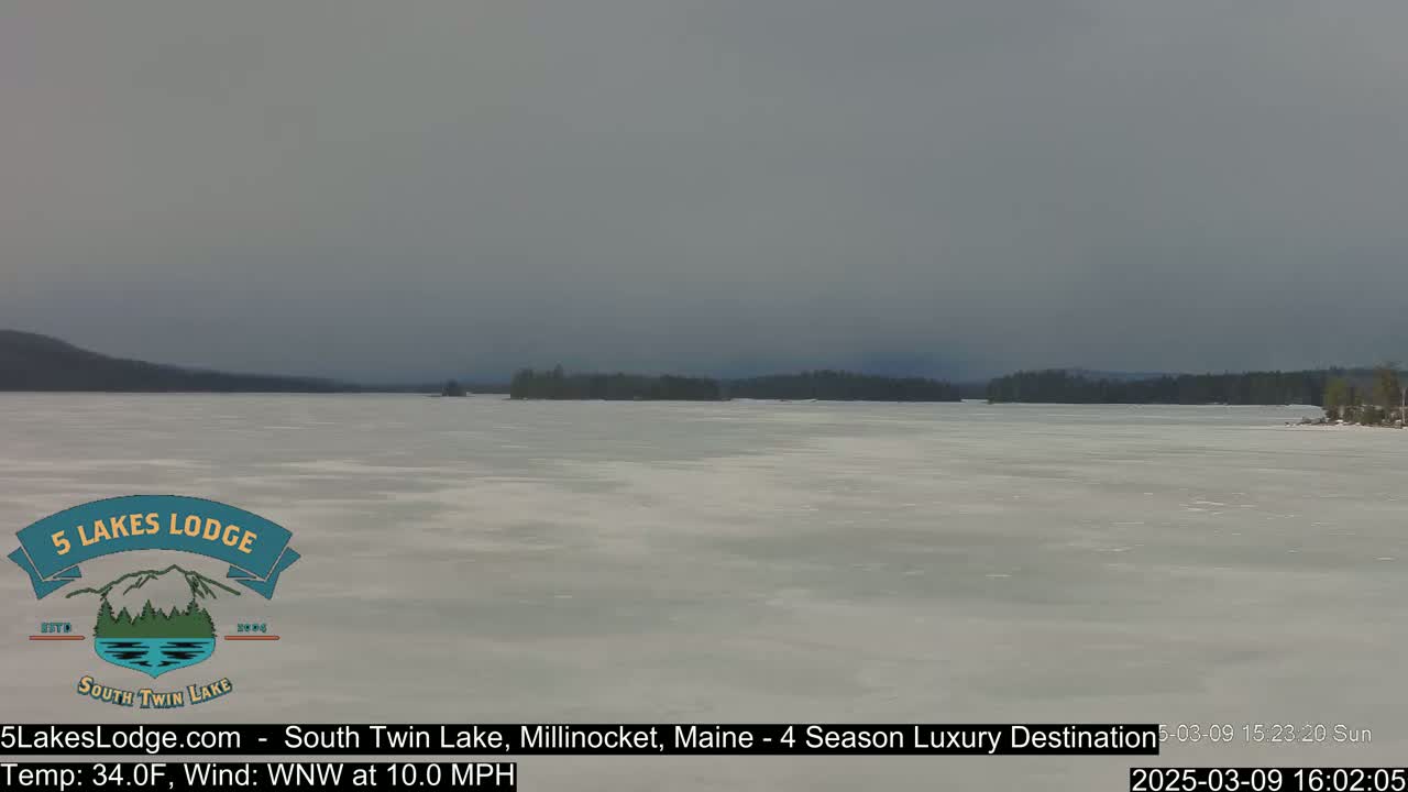 A mostly frozen lake stretches to a dark, tree-lined shore under an overcast sky.