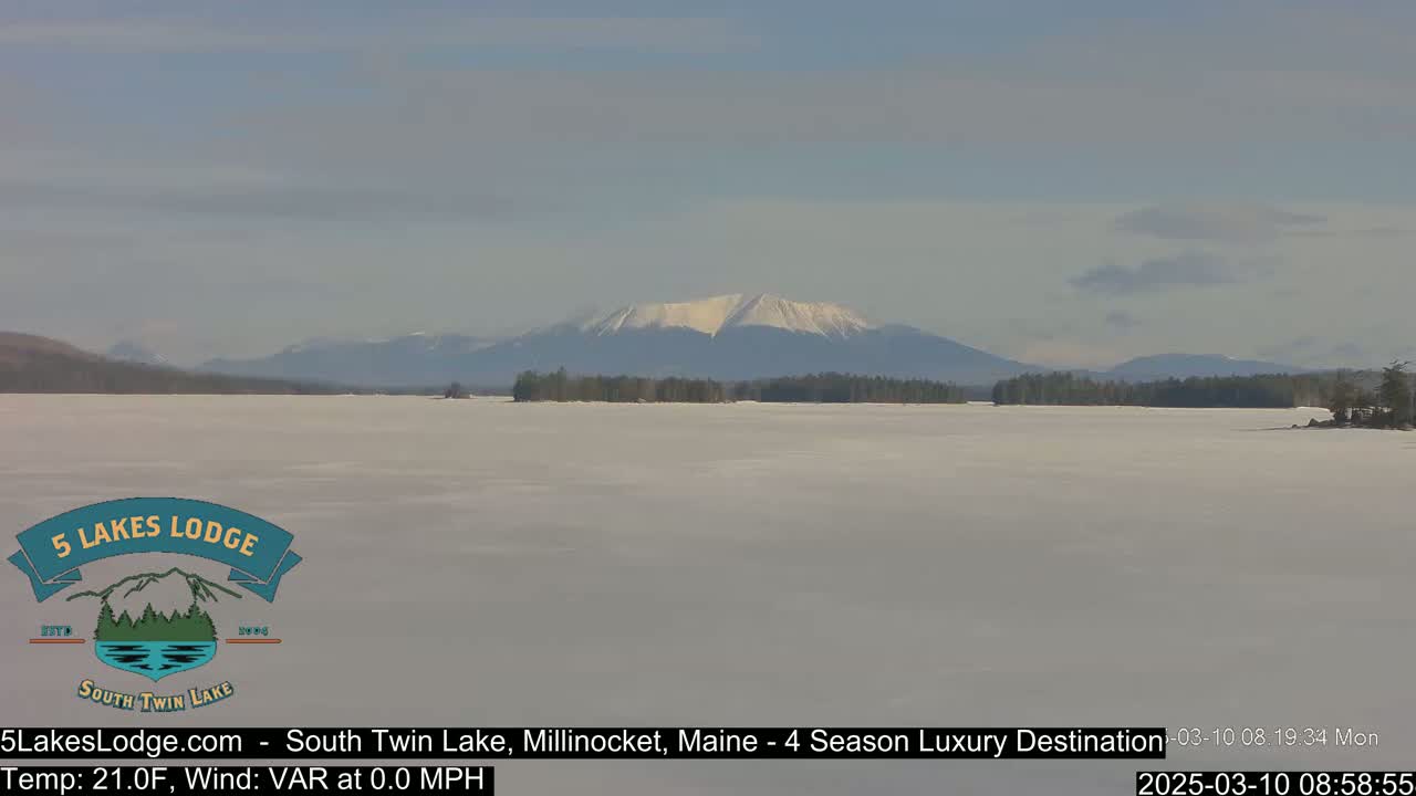 A snow-covered lake stretches to a distant, snow-capped mountain range under a mostly clear sky.