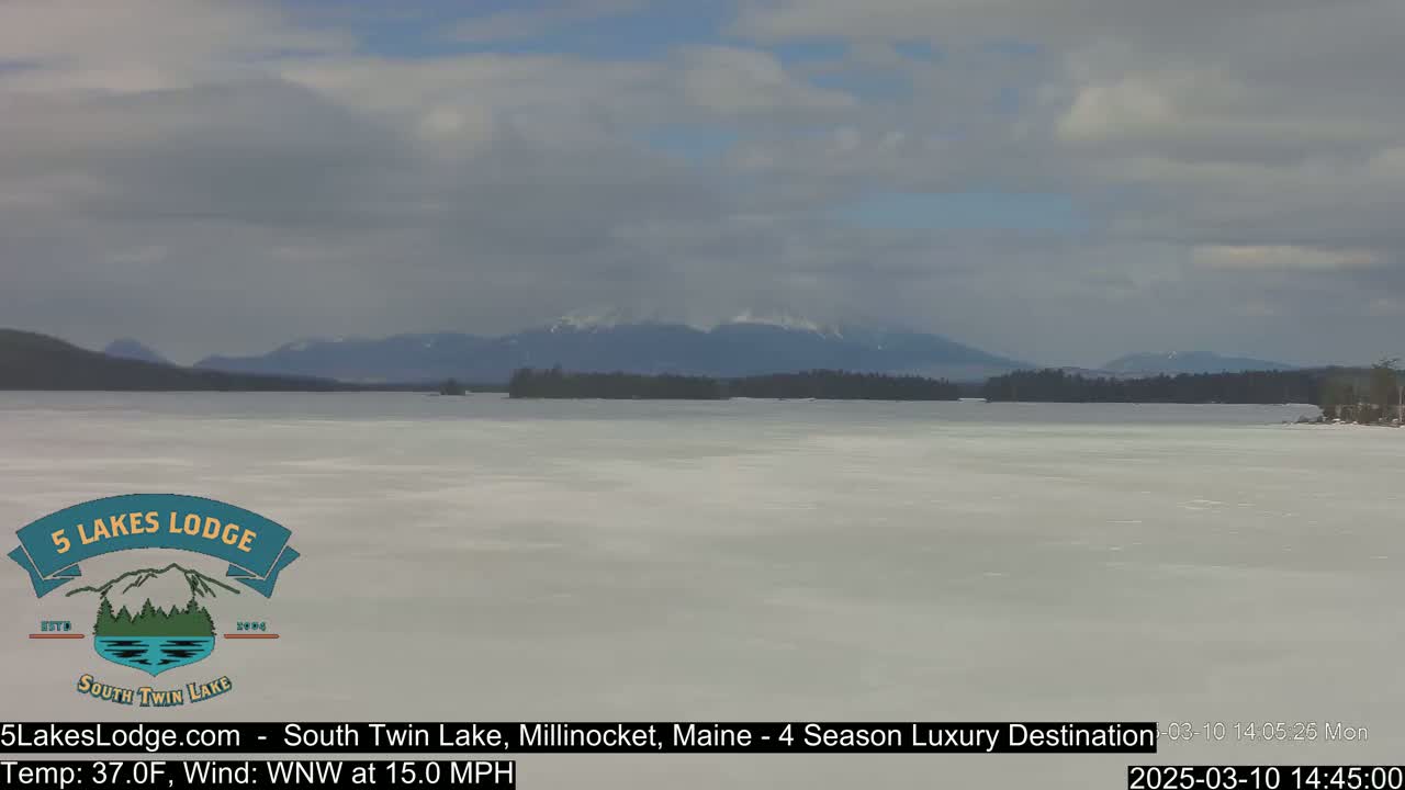 A mostly cloudy day reveals a frozen lake stretching towards snow-capped mountains under a pale sky with a 15 mph wind.