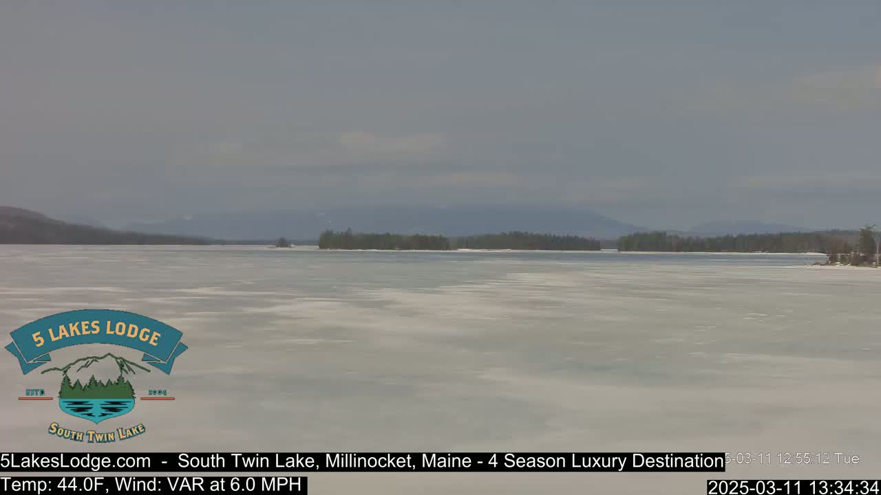 A mostly frozen lake stretches to a distant, tree-lined shoreline under a hazy sky, with a light wind.