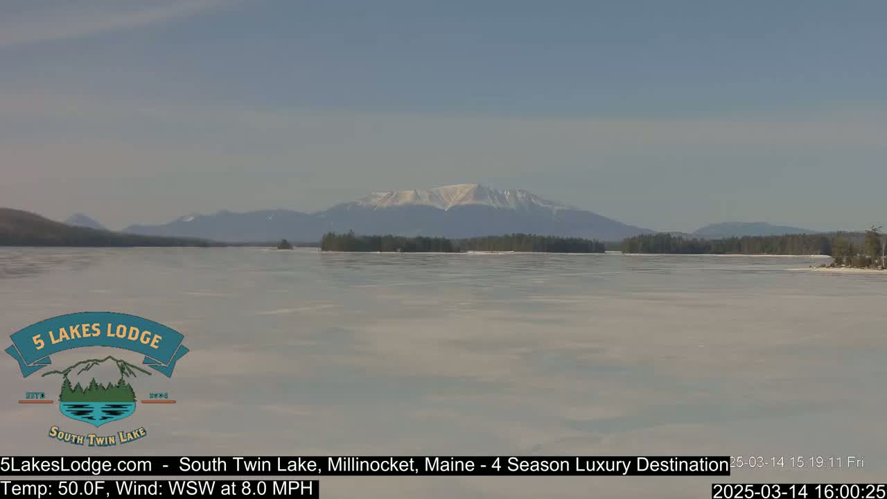 A partially frozen lake stretches towards snow-capped mountains under a clear sky.
