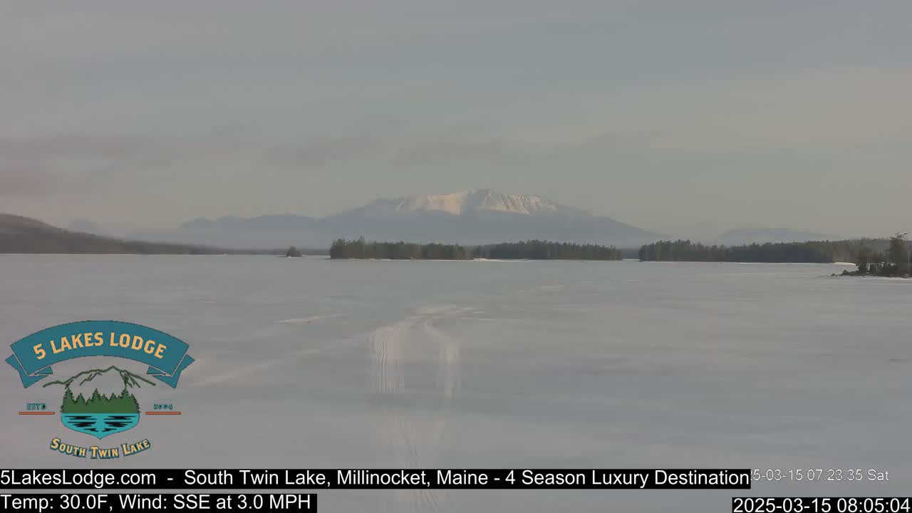 A snow-covered lake stretches toward distant mountains under a hazy sky.