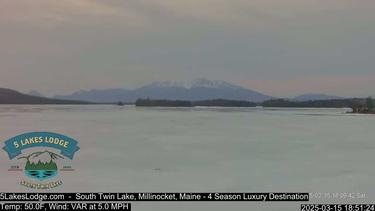 A partially frozen lake stretches toward snow-capped mountains under an overcast sky.