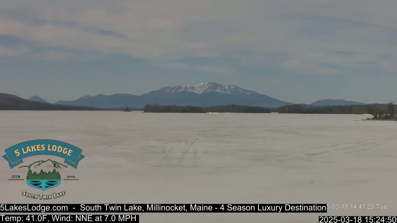 A snow-covered lake stretches to a distant mountain range with a partially snow-capped peak under a partly cloudy sky.