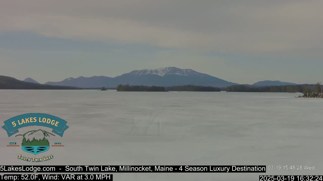 A partly frozen lake stretches to a snow-capped mountain range under a mostly cloudy sky.