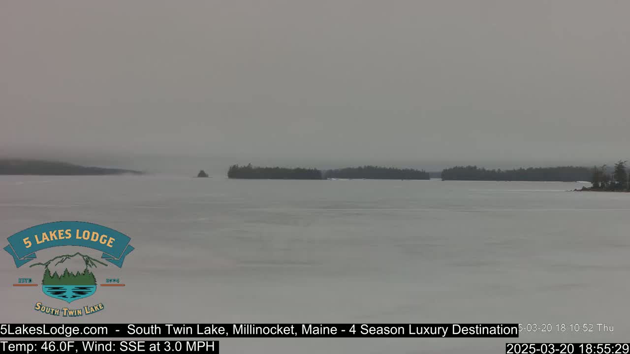 A mostly frozen lake is visible under an overcast sky, with dark tree lines on the far shore and small islands in the middle distance.