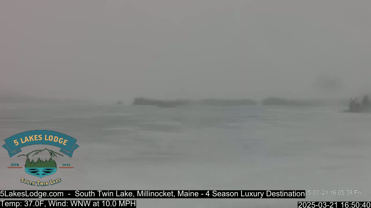 A snow-covered lake is visible under heavy snowfall with a strong wind.