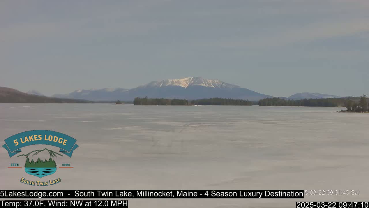 A partially frozen lake stretches towards snow-capped mountains under a mostly clear sky.