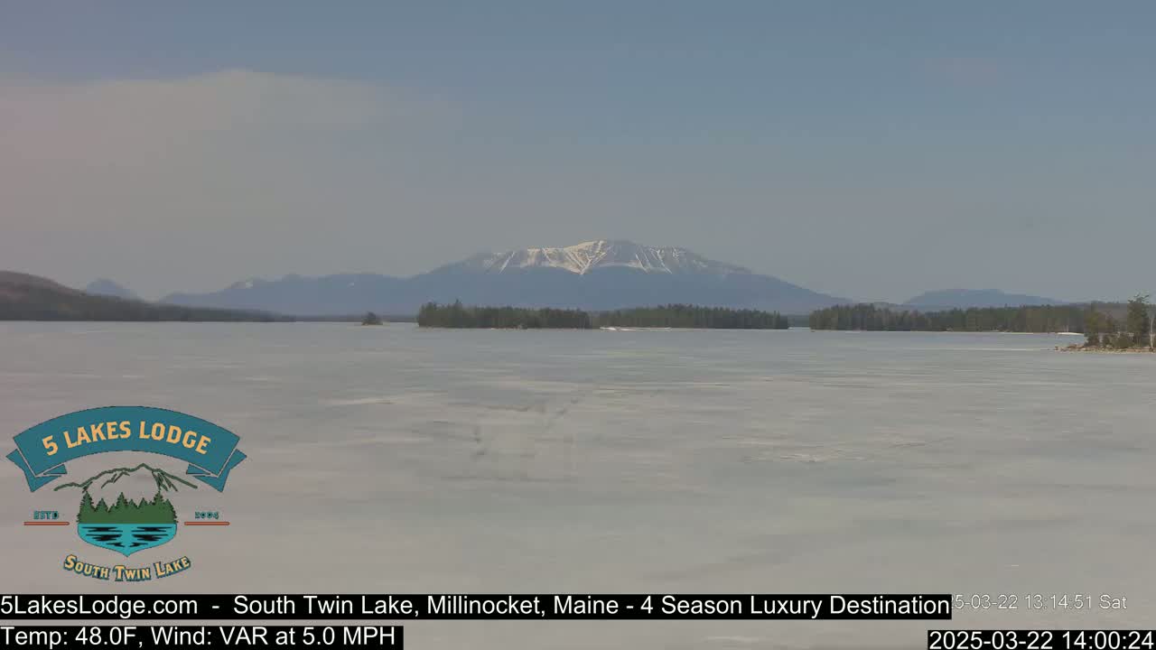 A partially frozen lake stretches towards snow-capped mountains under a clear sky.
