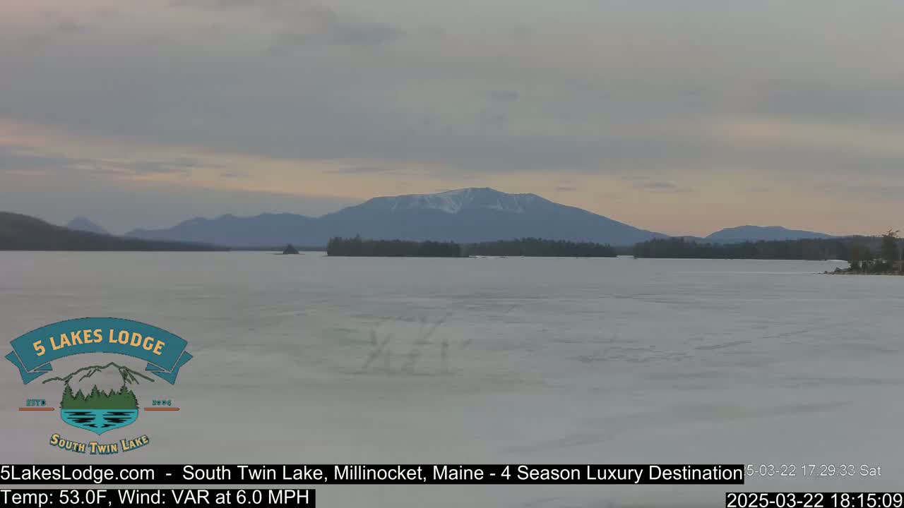 A partly frozen lake stretches towards a mountain range under a cloudy, dusky sky.